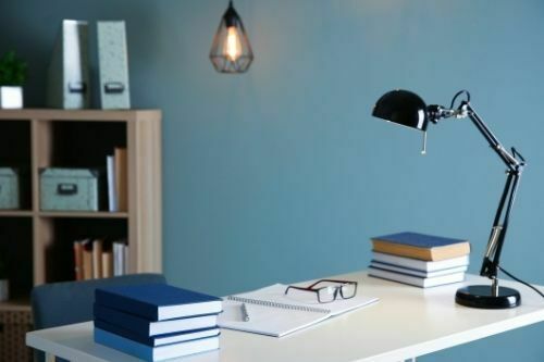 A lamp light kept on table in study room, also there are few books as well as the glassed are kept on open notebook on the table.