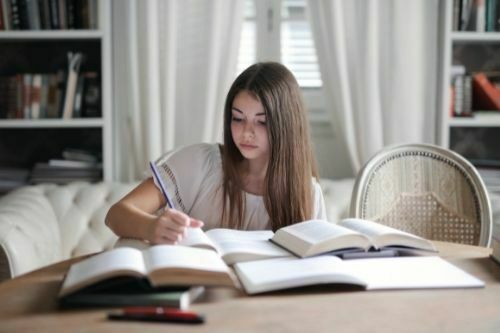a focused girl preparing for UPSC to become IPS office sitting on couch and 4 books are opened next on table. She is holding blue pen in her right hand.